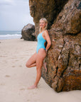 JAYD Swimwear model posing on a sandy beach next to a large rock, wearing the light blue Mykonos one-piece Tallow swimsuit.