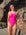 JAYD Swimwear model Kiera wearing the bright pink sorbet Tallow one-piece swimsuit, standing on a sandy beach in front of large brown and tan rocks.