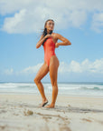 Athlete Lauren adjusting the fixed shoulder straps of the Blaze Zimmy one piece swimsuit whilst standing on the beach gazing off into the distance.