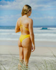JAYD Swimwear model Brodee wearing the yellow Ignite fixed-back, cheeky coverage Zimmy one piece swimsuit standing on a beach with ocean waves in the background.